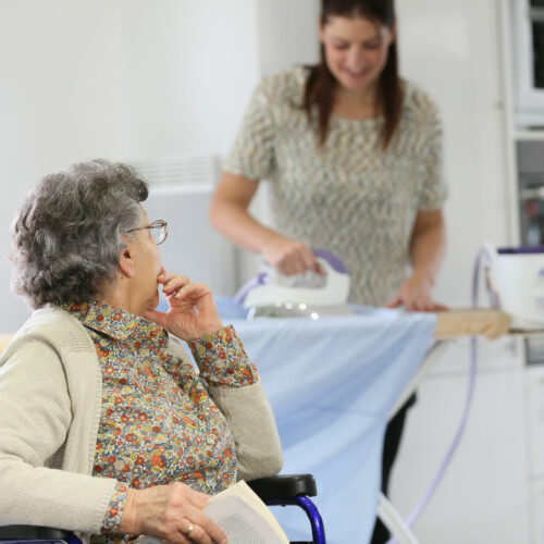 Elderly woman reading book while home helper irons laundry Elderly woman reading book while home helper irons laundry